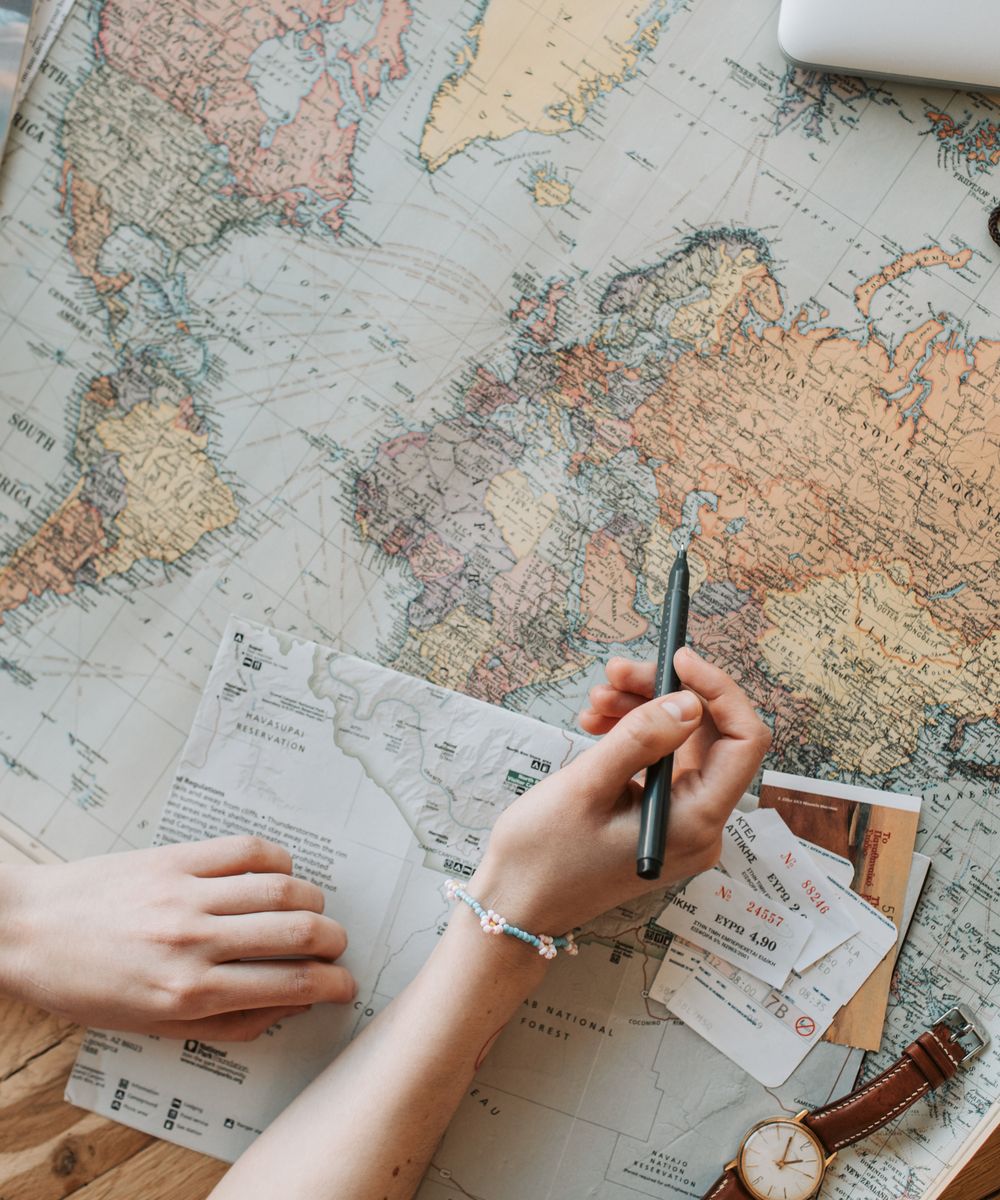 Hands marking a route on a printed map beside a camera and travel notebook.
